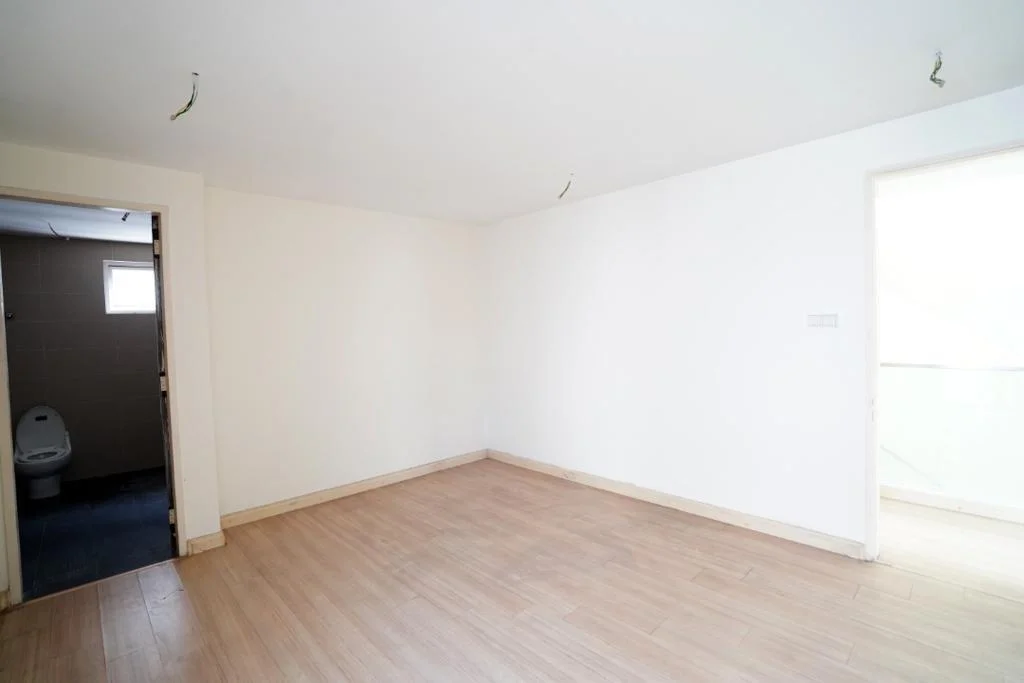 Room features: white painted walls, laminate flooring, doorway leading to bathroom, natural light from window, no visible furniture or decor, ceiling with exposed wiring, plain ceiling, neutral color scheme; style: minimalist.